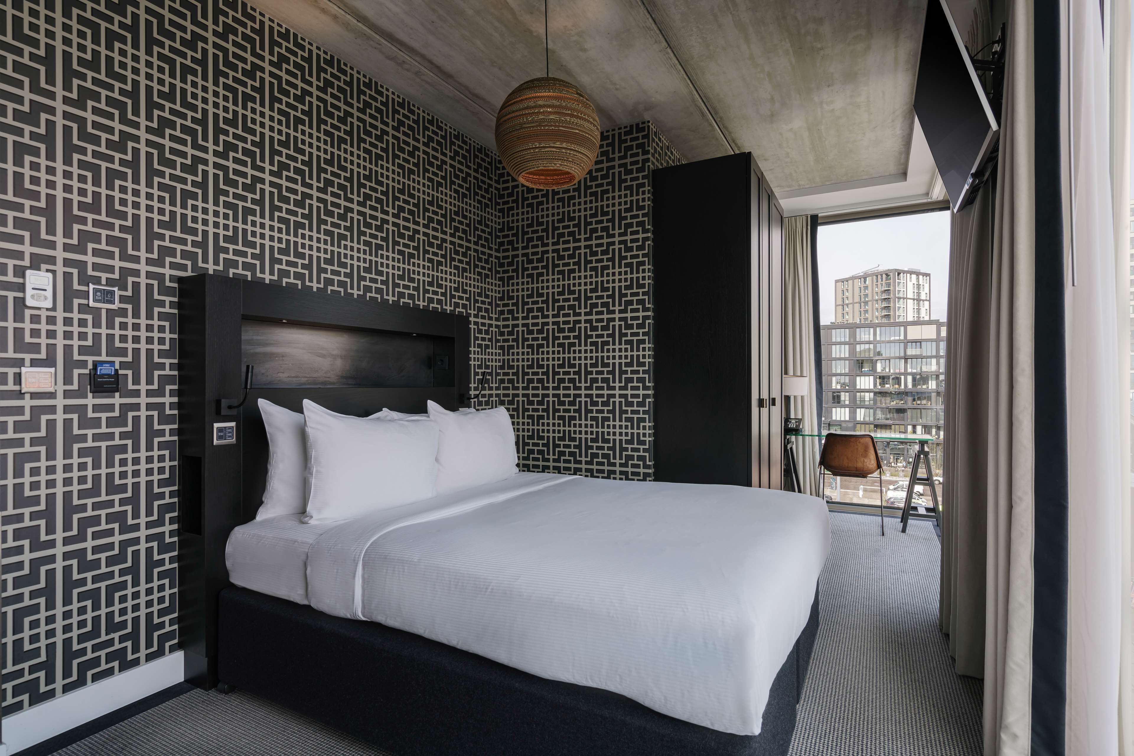 Une chambre d’hôtel moderne avec un grand lit, un papier peint à motifs, un luminaire suspendu et une vue sur la ville par la fenêtre.