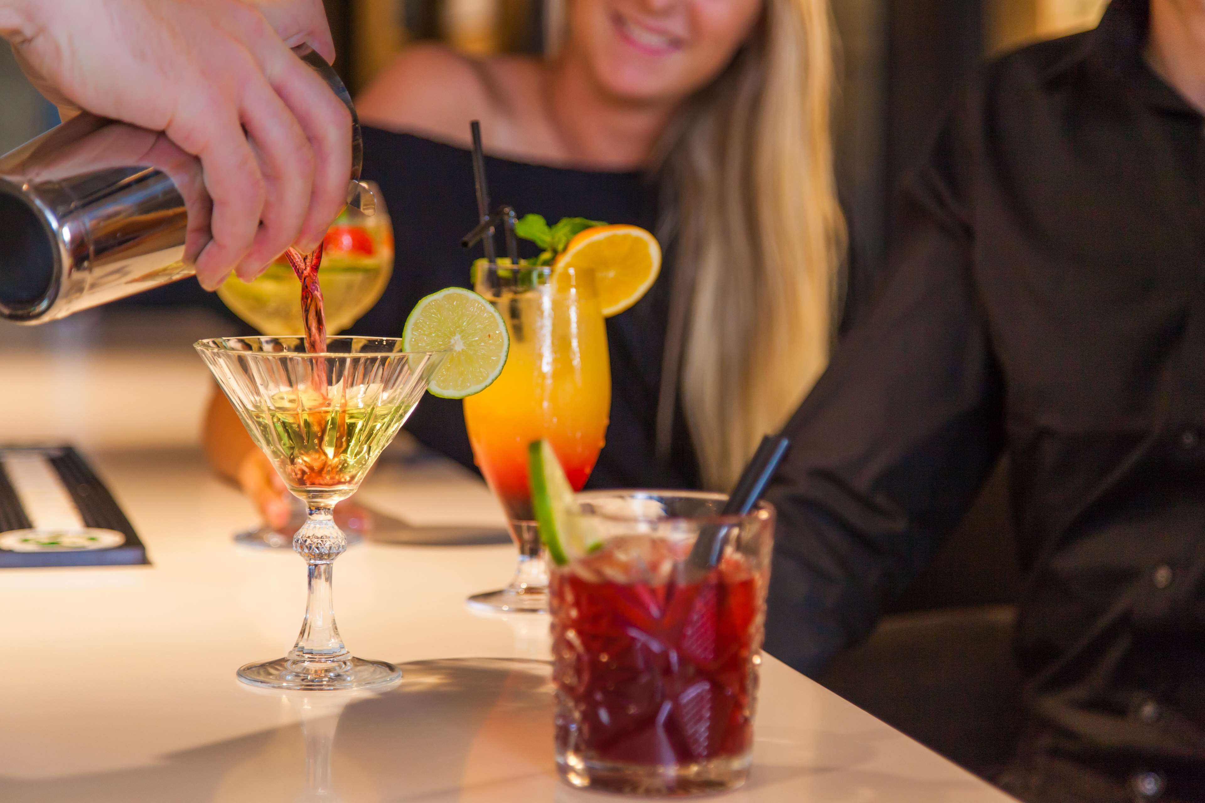 A bar setting with a person pouring a drink into a glass. There are three drinks on the counter, each with different garnishes.