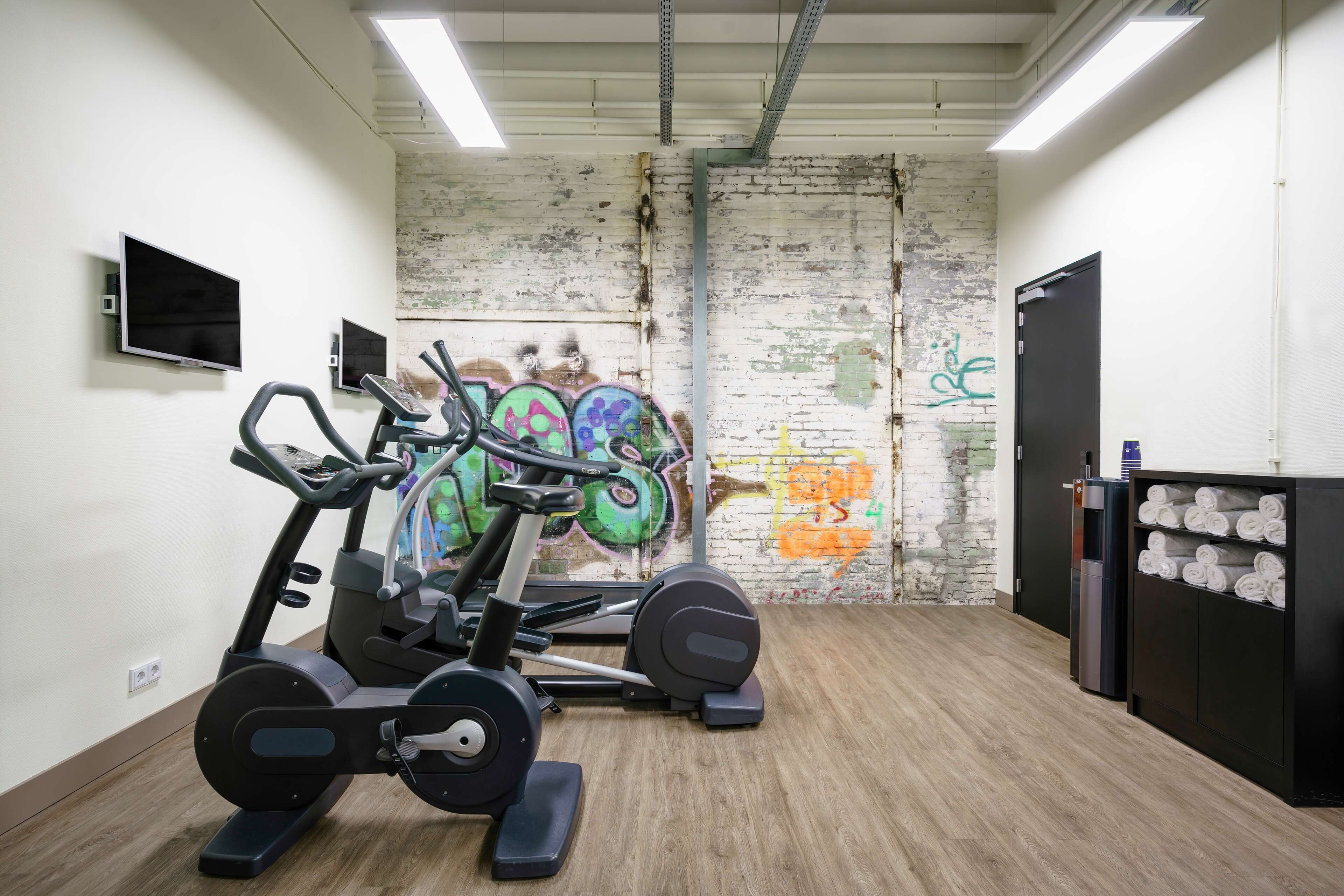Une salle de sport équipée de matériel d’exercice, un mur de briques couvert de graffitis, un meuble noir contenant des serviettes et un téléviseur fixé au mur.