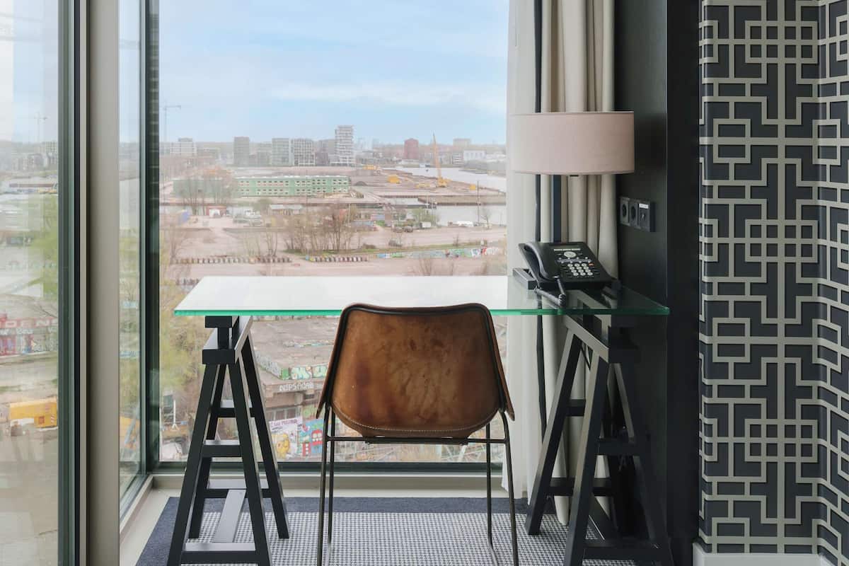 Une chambre d’hôtel avec une grande fenêtre, une table en verre, un fauteuil marron, une lampe et un mur à motifs.