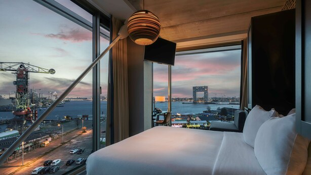 Habitación de hotel moderna con una cama grande, vista al paisaje urbano y una grúa de fondo.