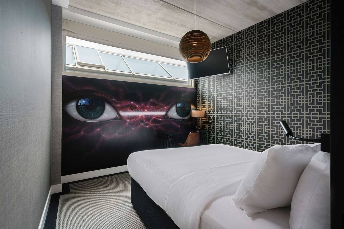 Une chambre d’hôtel moderne dotée d’un grand lit, d’un téléviseur fixé au mur et d’une fresque murale originale sur le thème de l’œil.