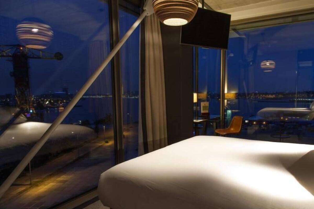 Une chambre d’hôtel avec une grande fenêtre, un lit, une chaise et une lampe moderne.