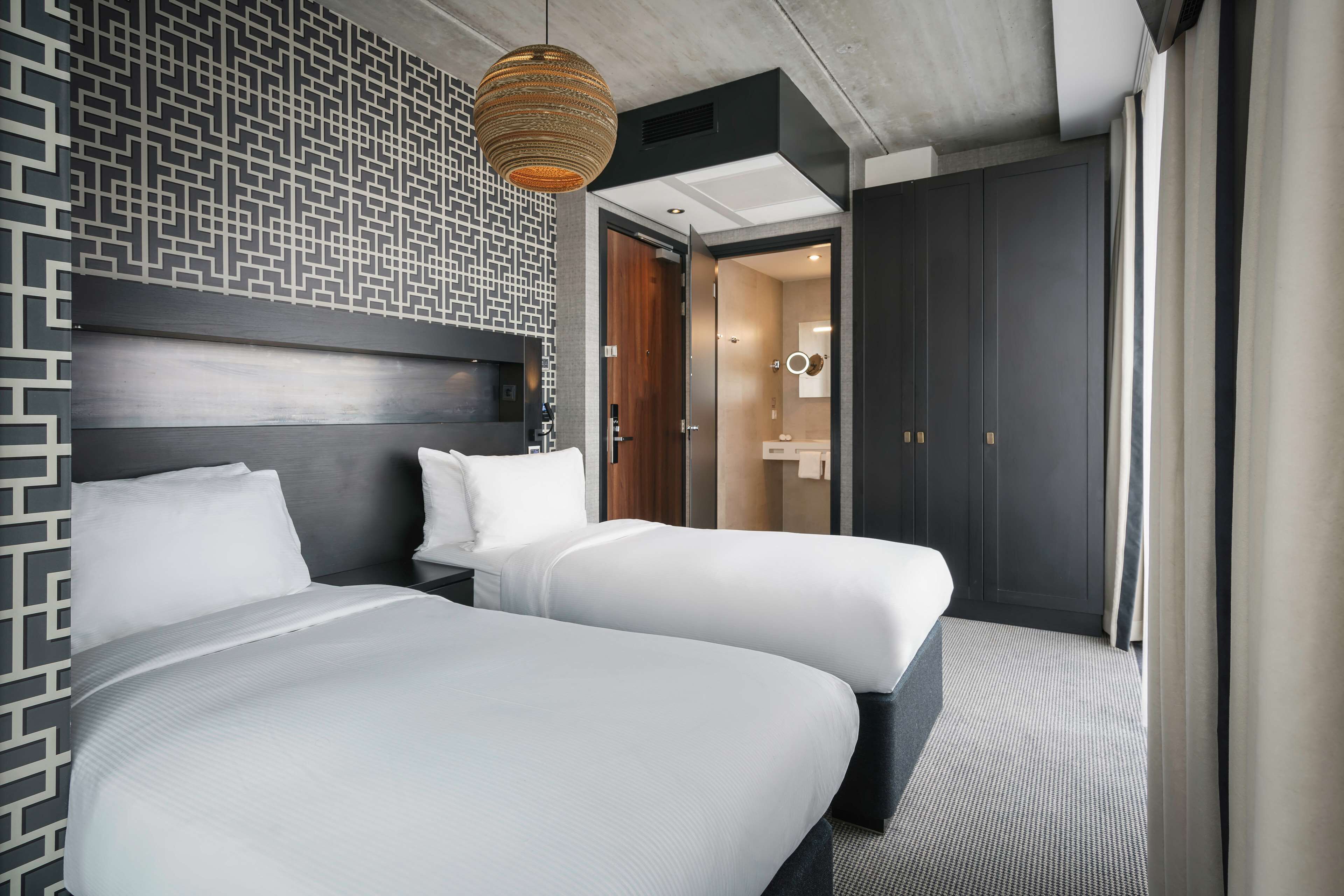 A modern hotel room with two beds, a patterned wall, a hanging light fixture, and a view of the bathroom.