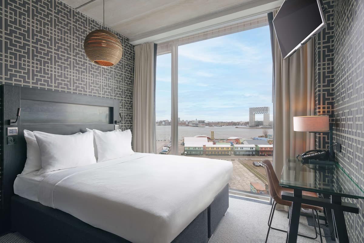 Une chambre d’hôtel avec un grand lit, un bureau avec une chaise, une télévision et une vue sur la ville grâce à de grandes fenêtres.