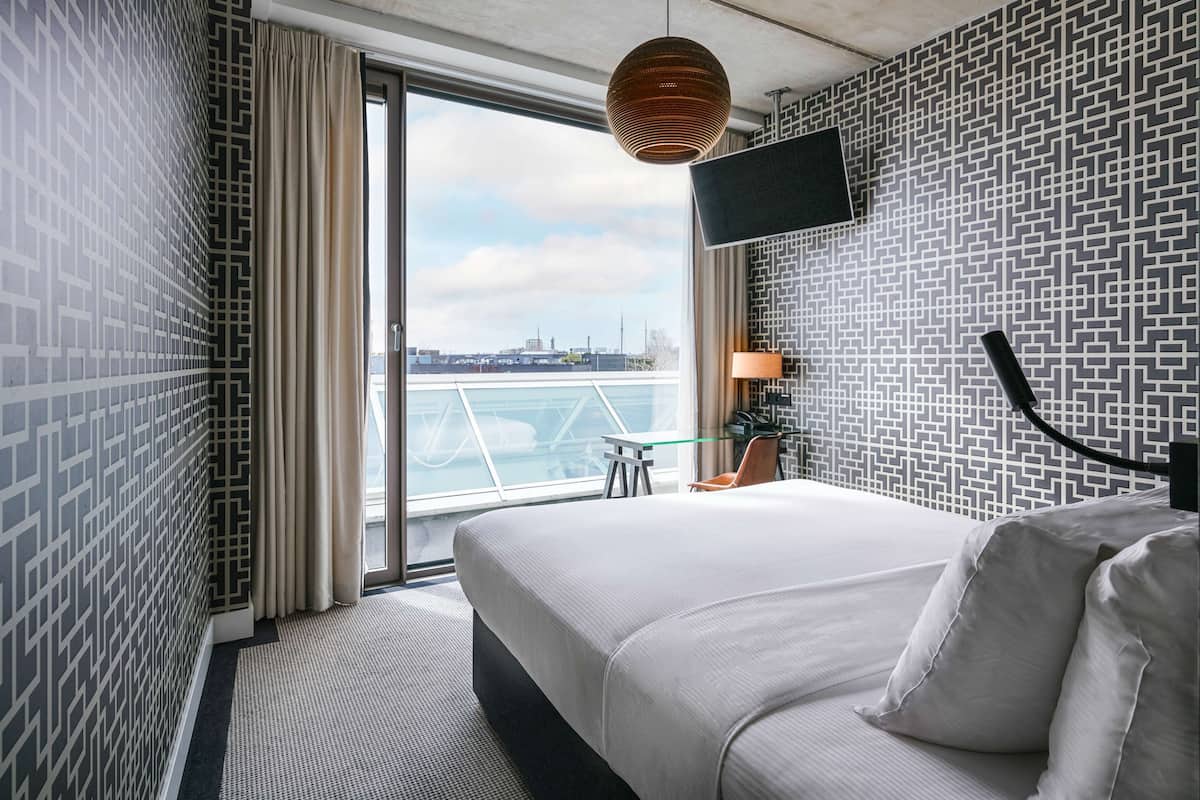 Une chambre d’hôtel moderne avec un grand lit, un téléviseur fixé au mur, une vue sur la ville par la fenêtre et un motif décoratif sur le mur.