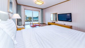 Hotellihuone, jossa on suuri sänky, televisio, työpöytä lampun kanssa ja parvekeovista avautuva näkymä merelle.