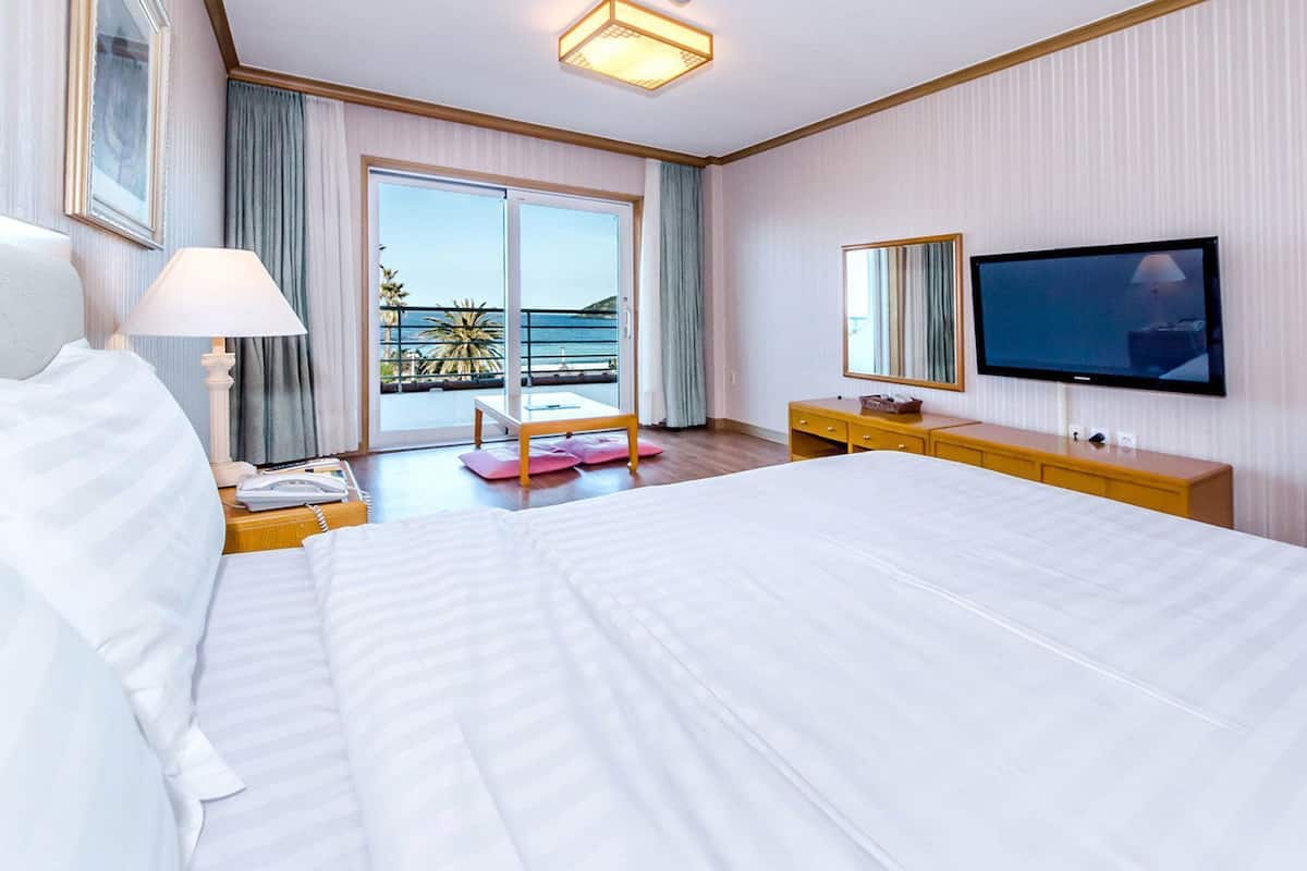 A hotel room with a large bed, a television, a desk with a lamp, and a view of the ocean through the balcony doors.