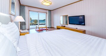 Habitación de hotel con una cama grande, un televisor, un escritorio con lámpara y vista al océano a través de las puertas del balcón.