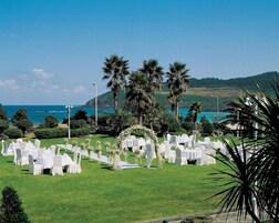 An outdoor wedding venue with white tables and chairs, a floral arch, and a view of the ocean and hills.