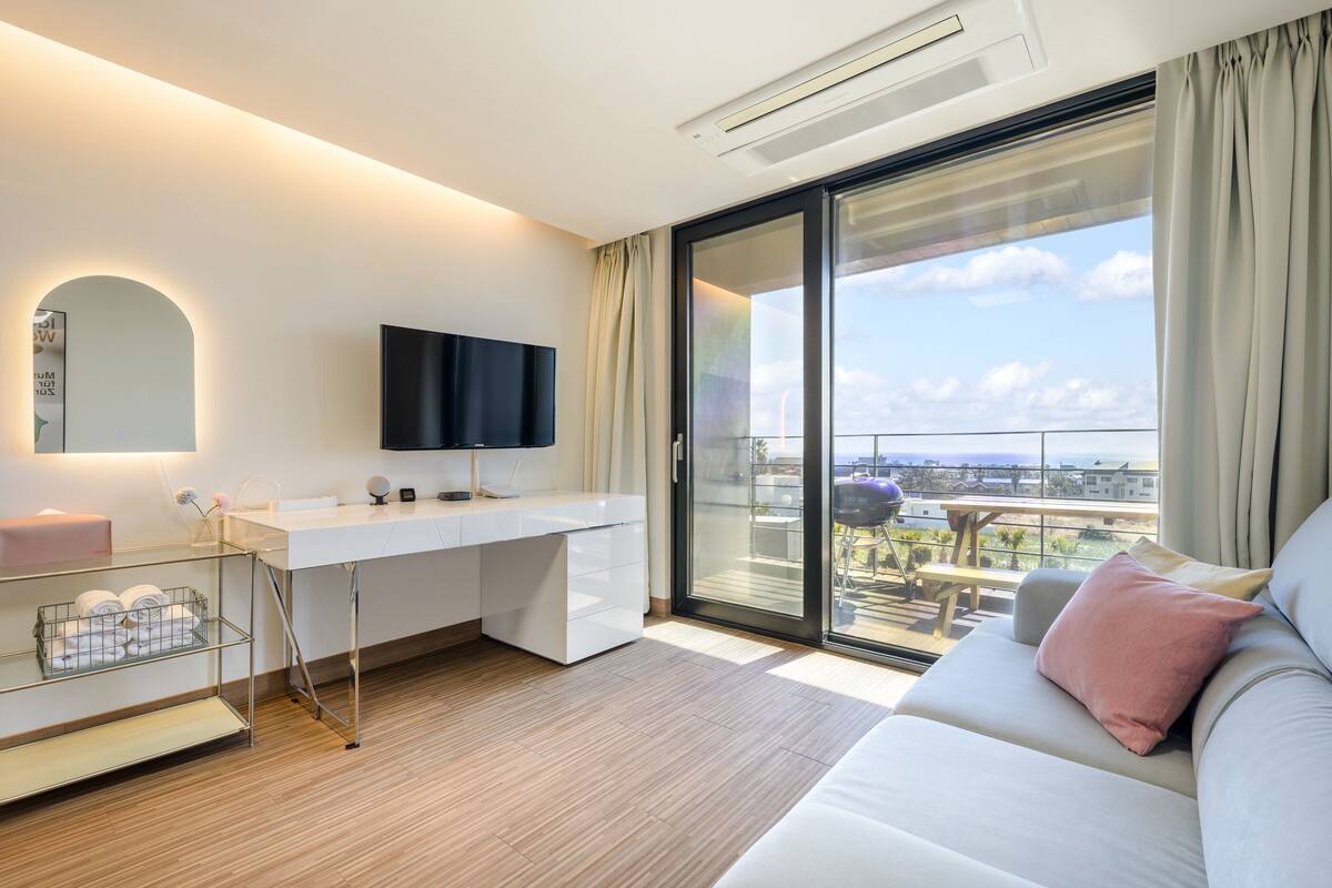 A modern hotel room with a large sliding glass door to a balcony, a white desk with a flat-screen TV, a sofa with cushions, and a small table with chairs.