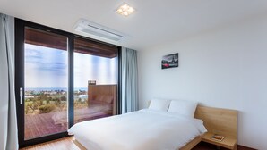 A bedroom with a large bed, a wooden headboard, a nightstand, a painting, and a view through a sliding glass door. - IRO Jeju (Seogwipo)