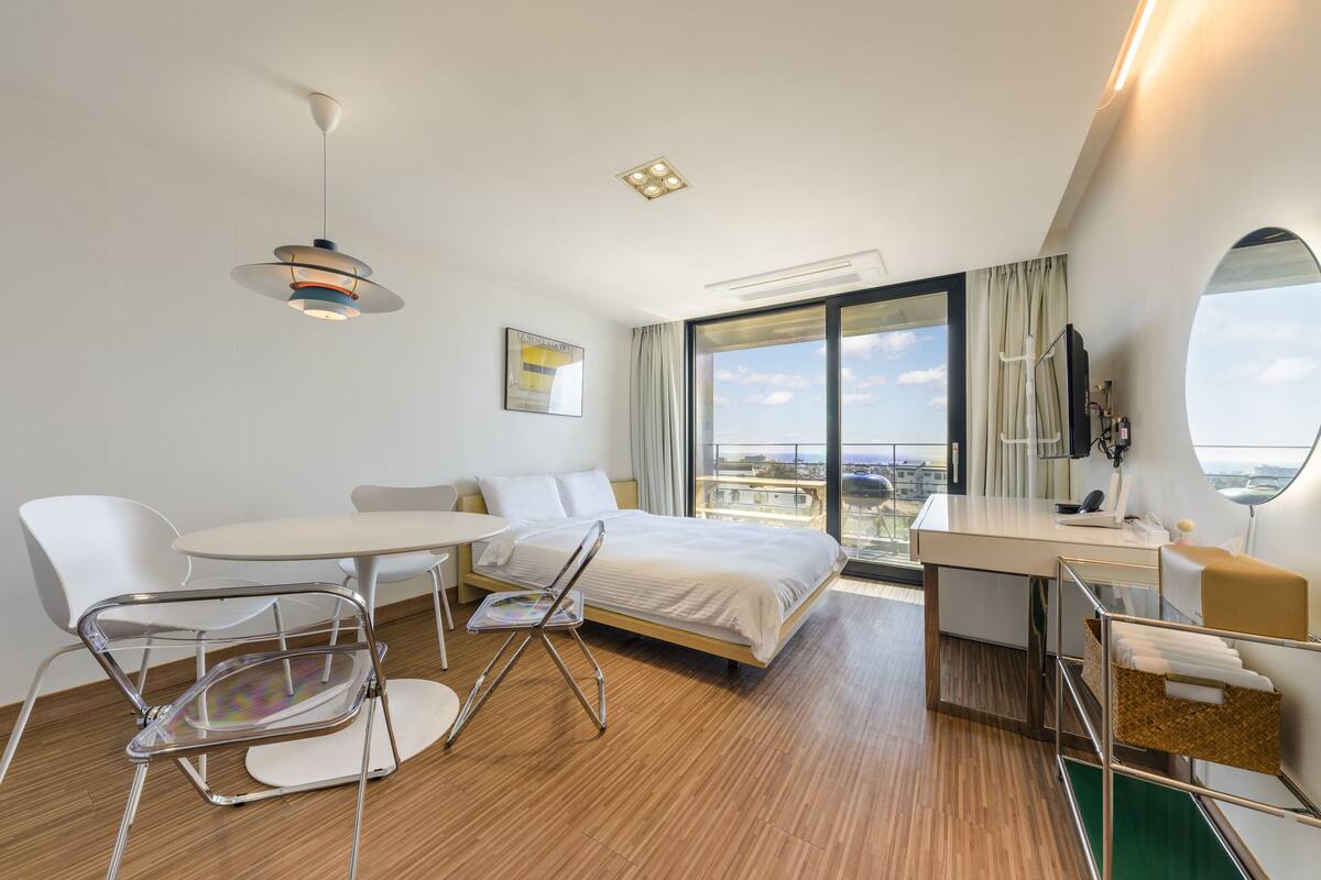 A modern hotel room with a bed, a desk, a chair, a table, and a large window offering a city view.