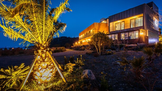 A modern building with a flat roof and large windows, surrounded by a landscaped area with palm trees and rocks. - IRO Jeju (Seogwipo)