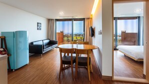 A hotel room with a wooden floor, a bed, a sofa, a dining table with chairs, a TV, and a view of the city. - IRO Jeju (Seogwipo)
