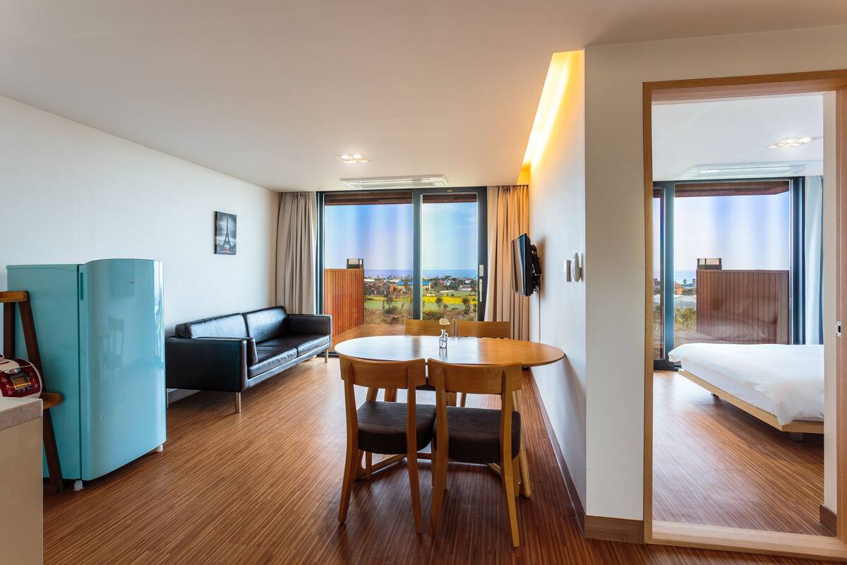 A hotel room with a wooden floor, a bed, a sofa, a dining table with chairs, a TV, and a view of the city.