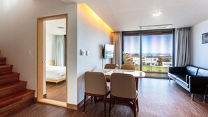 A hotel room with a wooden staircase, a dining area with a table and chairs, a black leather sofa, and a large window with a view of buildings and greenery. - IRO Jeju (Seogwipo)