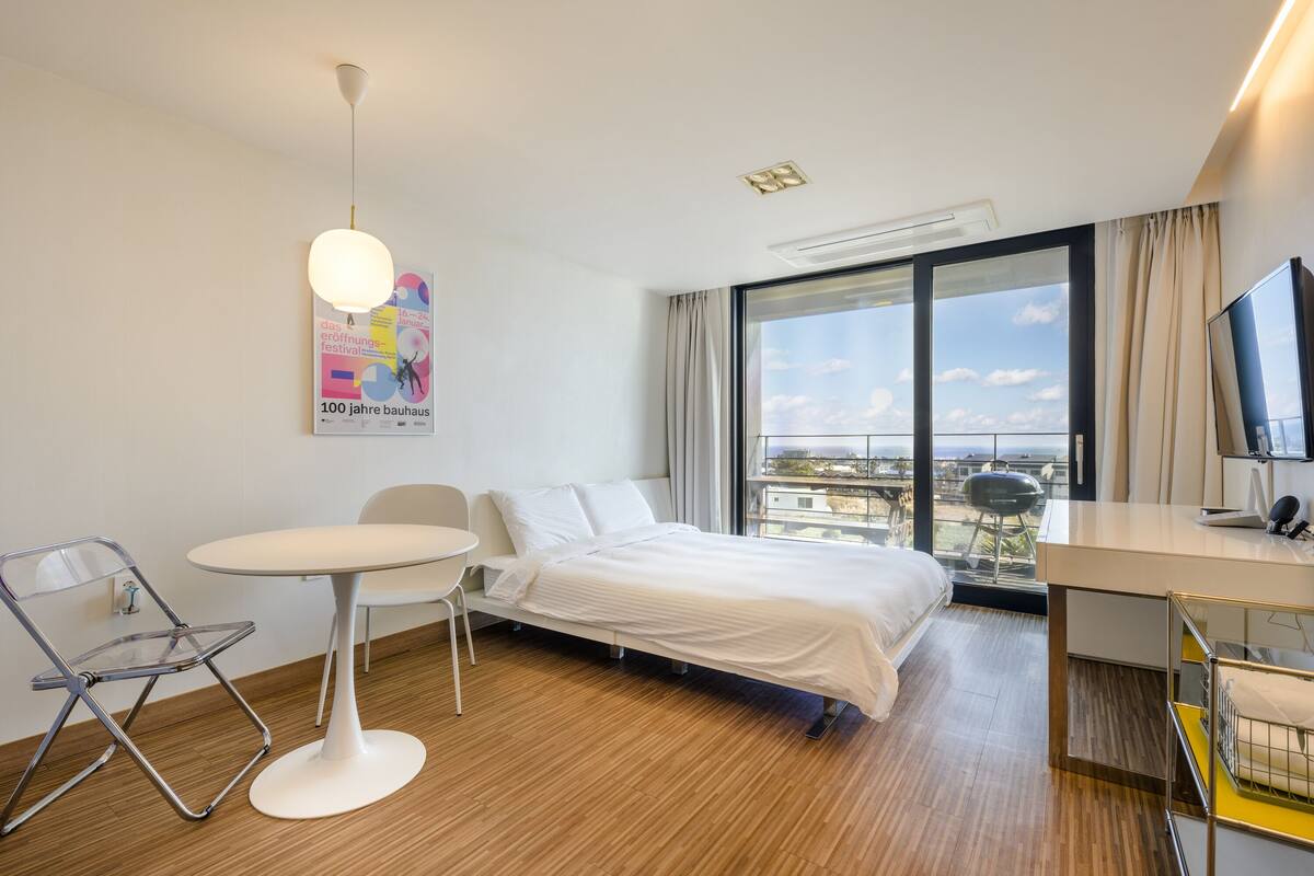 A modern hotel room with a bed, a desk, a chair, and a large window offering a city view.