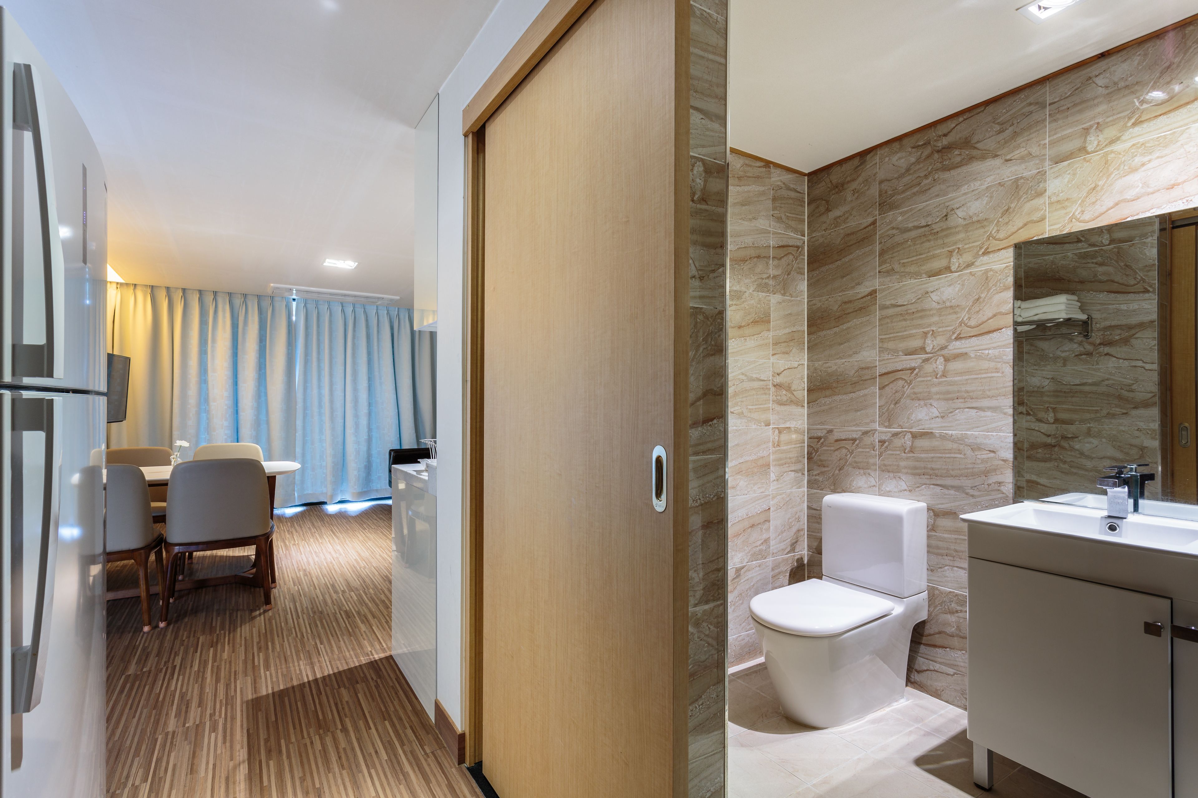 A modern hotel room with a wooden door, a dining area with a table and chairs, and a bathroom with a marble wall and white fixtures.