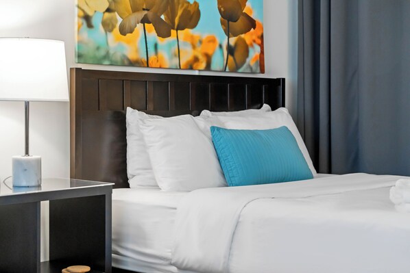 A neatly made bed with white and blue bedding, a modern bedside table with a lamp, and a wall-mounted painting of flowers. - BCA Furnished Apartments (Atlanta)