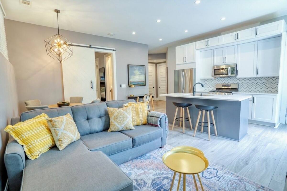 Een moderne woonkamer met een grijze bank, gele kussens met een patroon, een blauw vloerkleed en een gele poef.