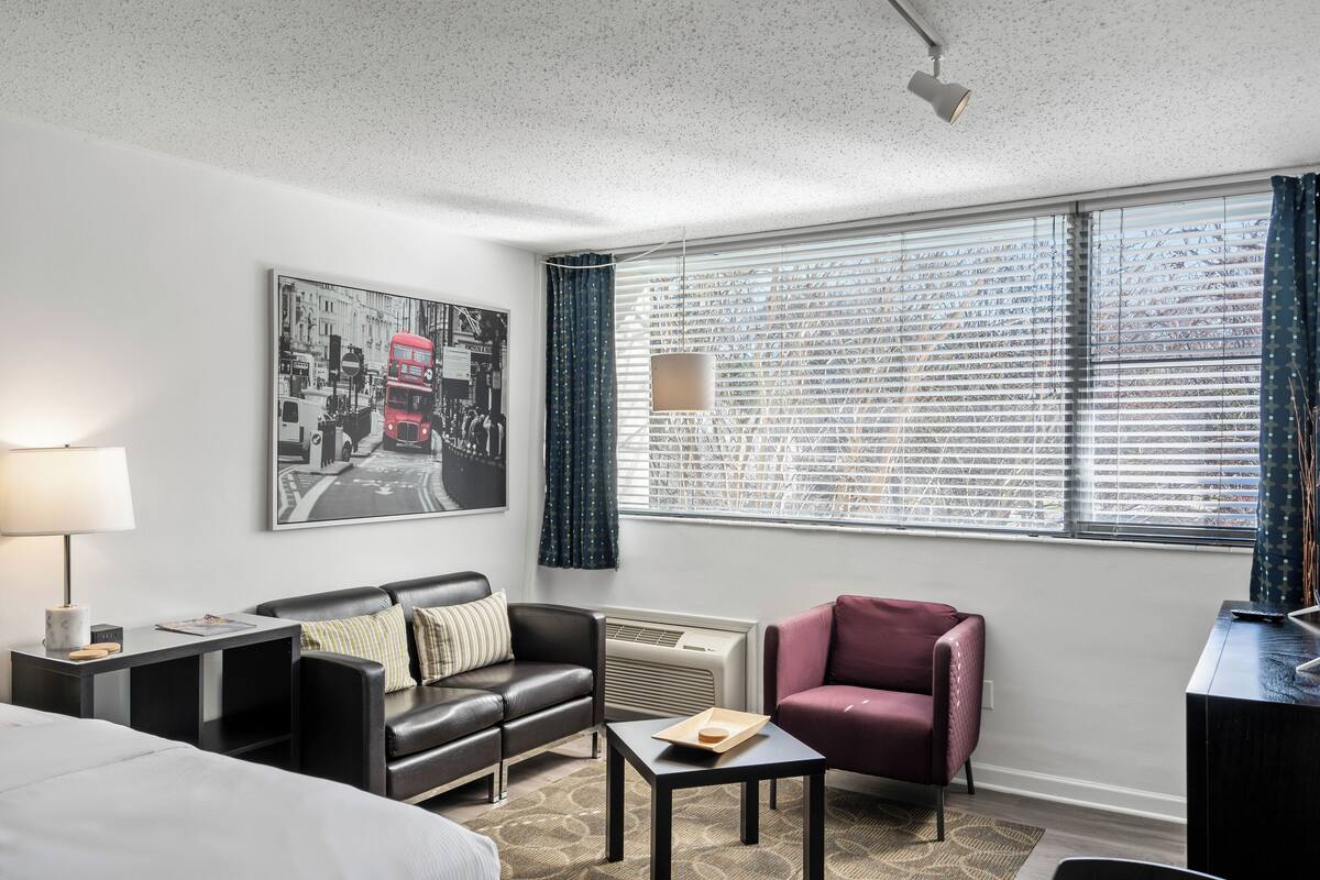 Een hotelkamer met een bed, een zwarte leren bank, een kleine tafel, een bureau, een lamp, een groot raam met jaloezieën en een stadsgezichtposter aan de muur.