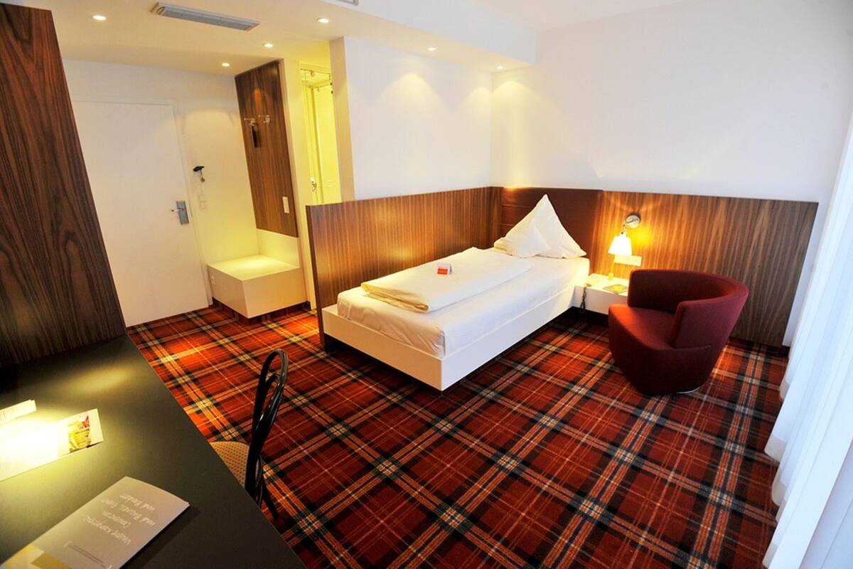 A hotel room with a bed, a red chair, a small table, and a plaid carpet.