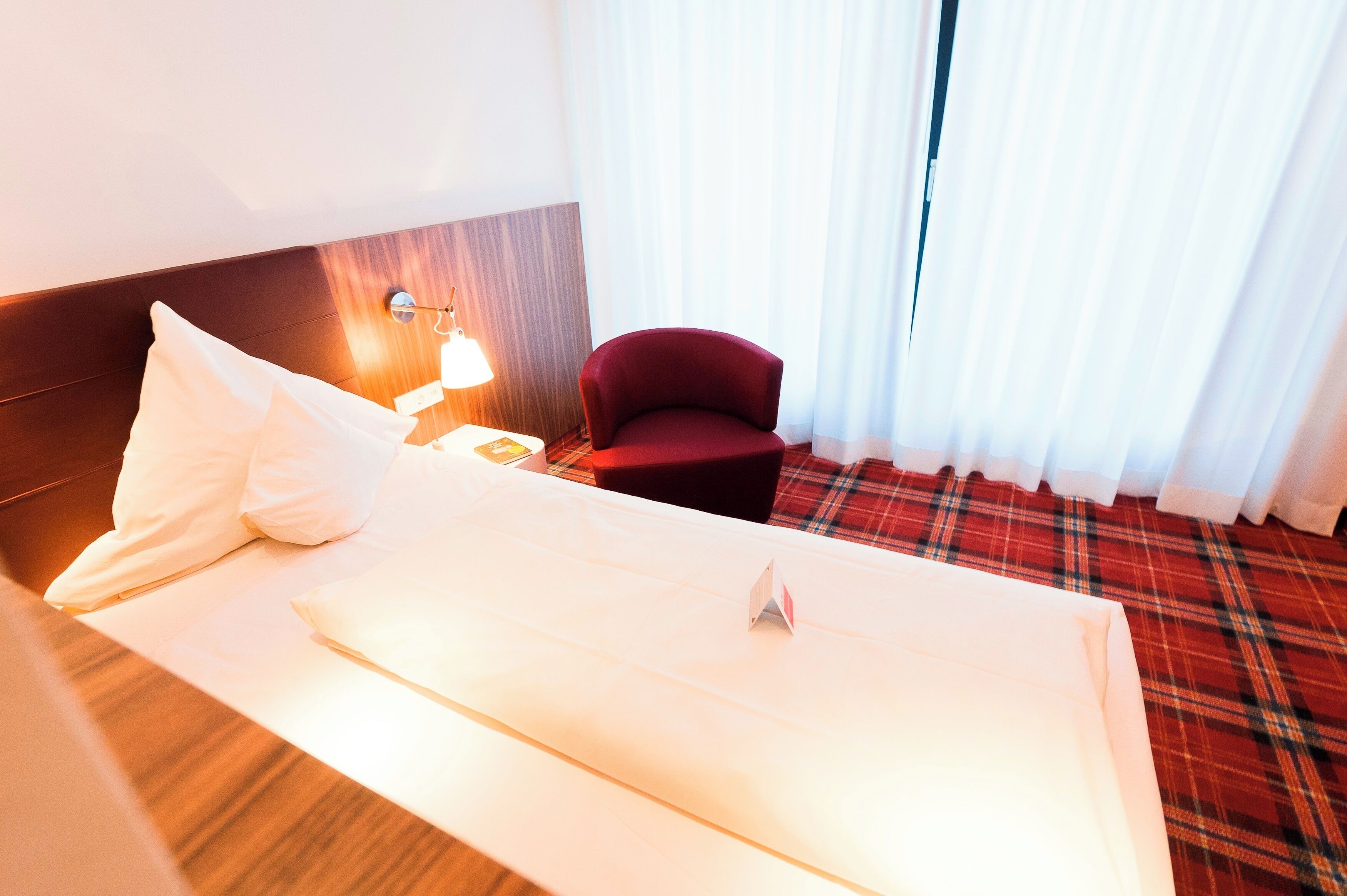 A hotel room with a bed, a red armchair, a nightstand with a lamp, and a window with curtains.