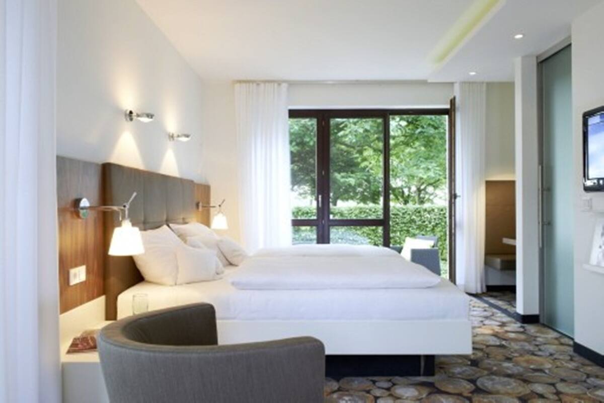 A modern hotel room with a large bed, a seating area, and a view of greenery outside.