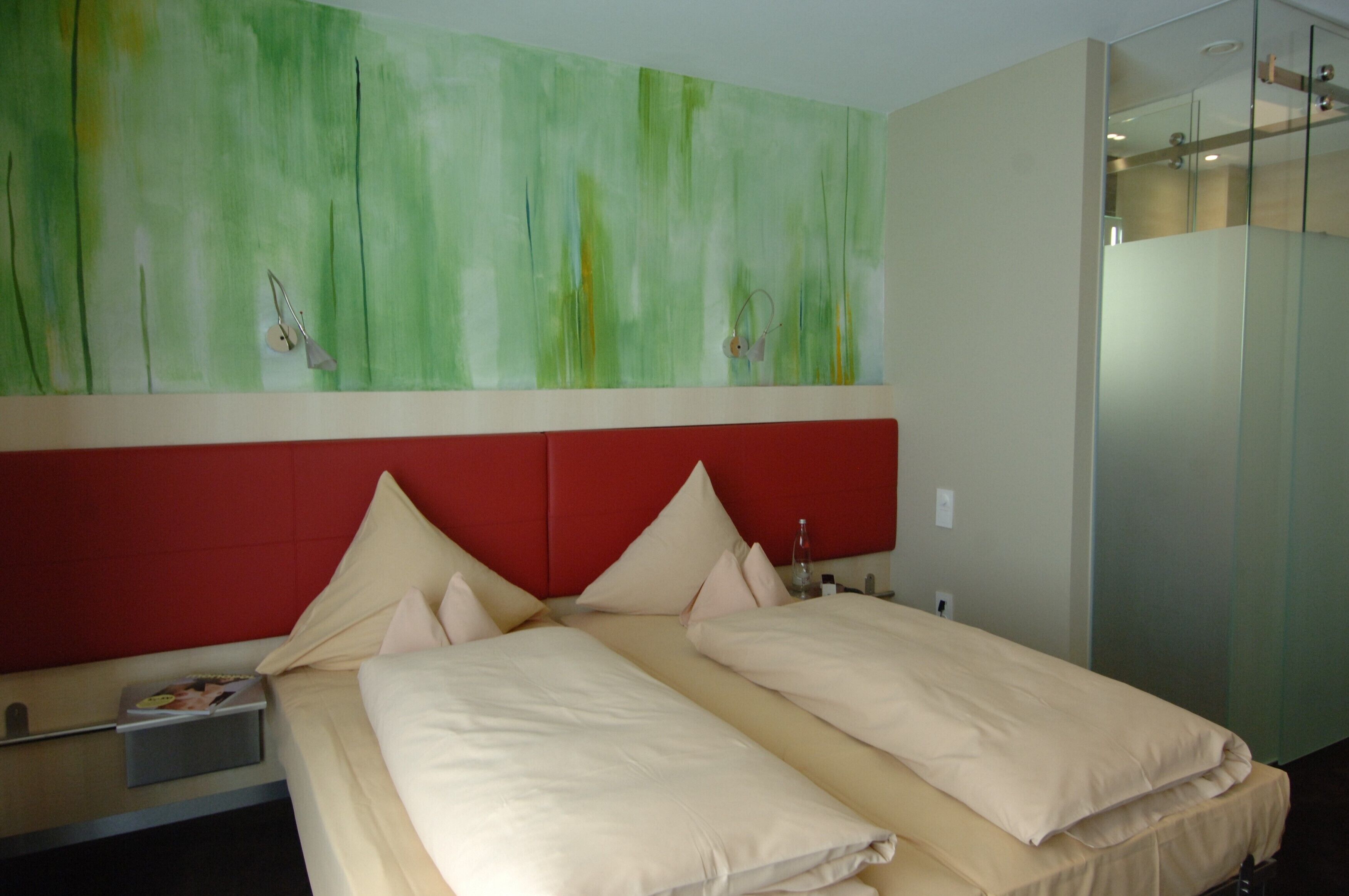 Ein Hotelzimmer mit Bett, Nachttisch, dunkelrotem Kopfteil und einem grünen abstrakten Gemälde an der Wand.