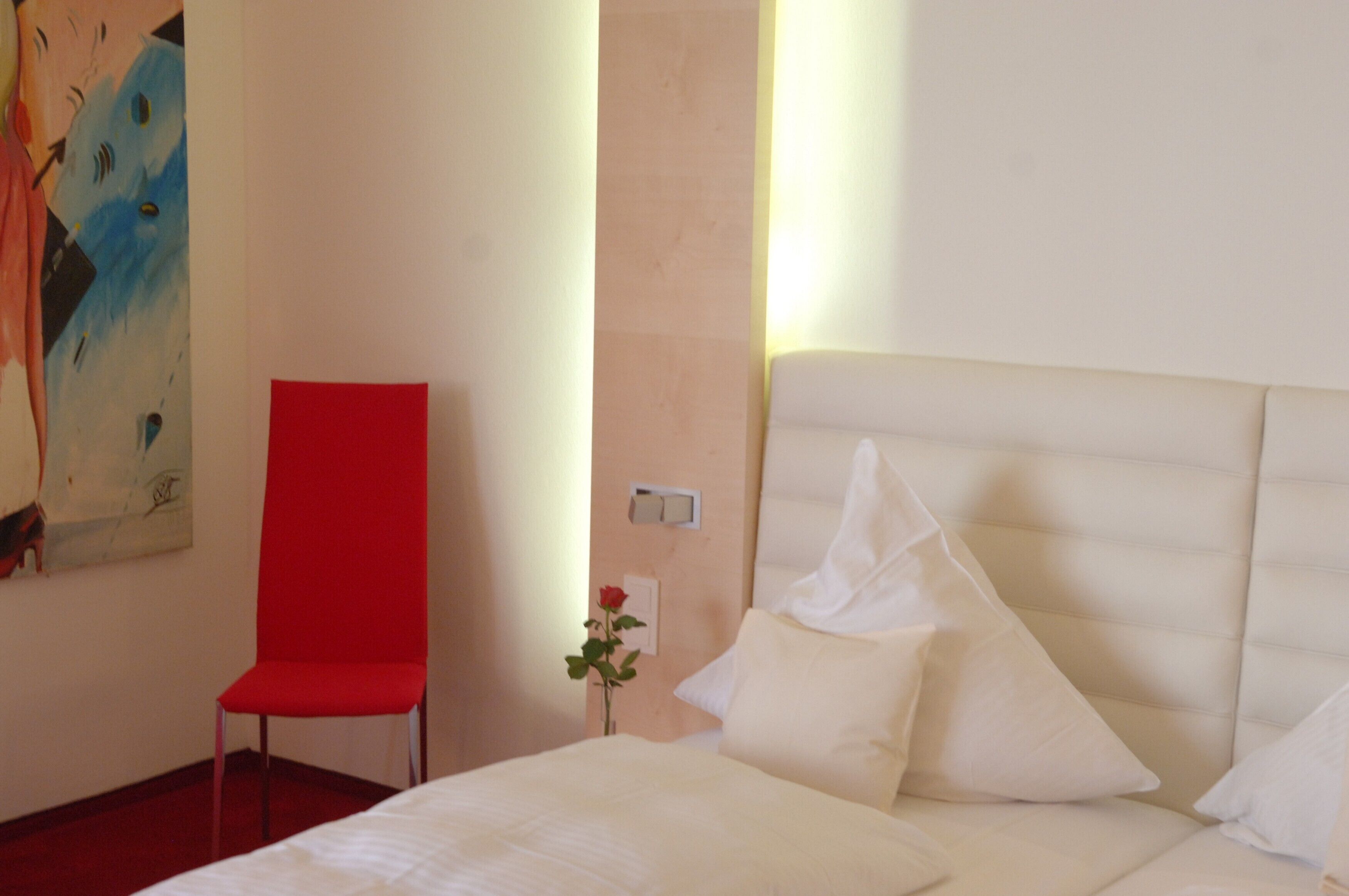 Ein Hotelzimmer mit einem Bett, einem roten Sessel, einem Gemälde an der Wand und einer Vase mit einer Rose.