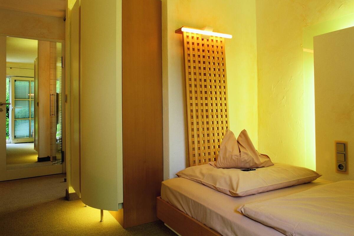 A hotel room with a bed, a wooden wardrobe, a wall-mounted light fixture, and a door leading to a bathroom.