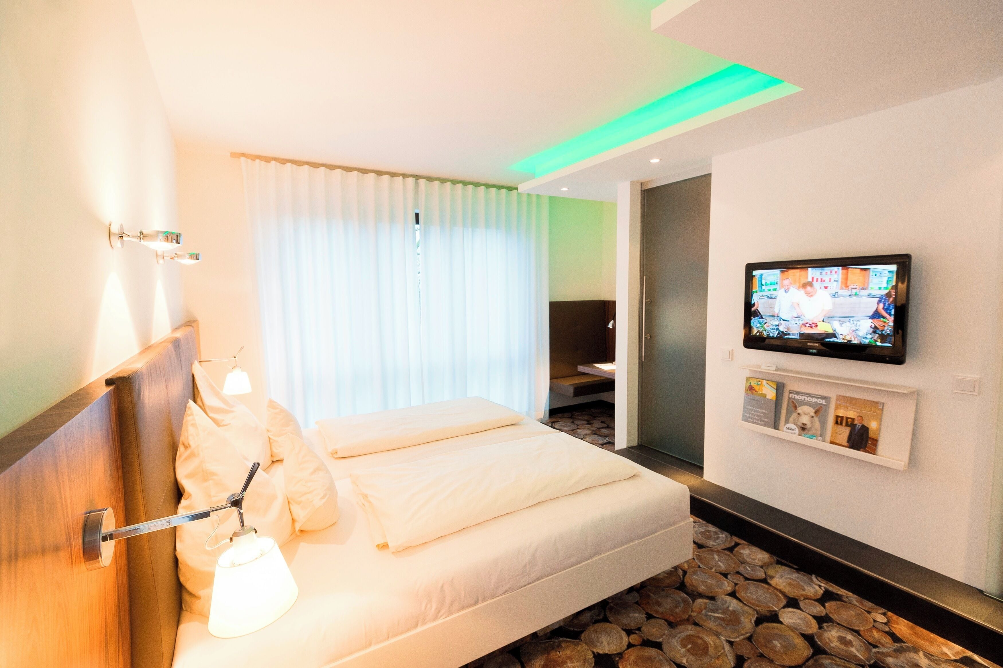 A hotel room with a bed, a TV, a wall-mounted shelf with pictures, and a window with curtains.