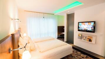 A hotel room with a bed, a TV, a wall-mounted shelf with pictures, and a window with curtains.