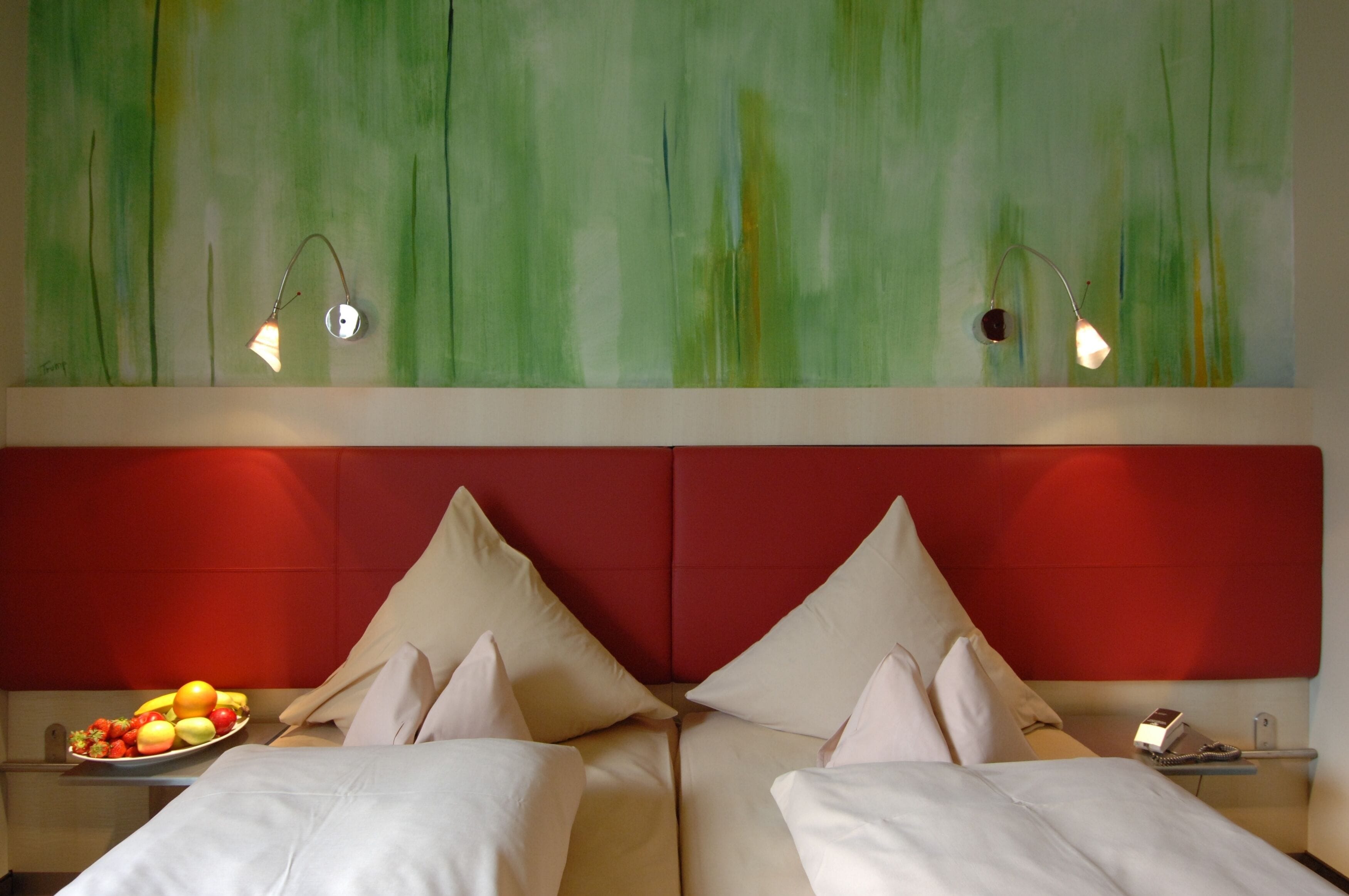 Ein Hotelzimmer mit zwei Betten, einer Obstschale auf dem Nachttisch und einem abstrakten Wandbild in Grün.
