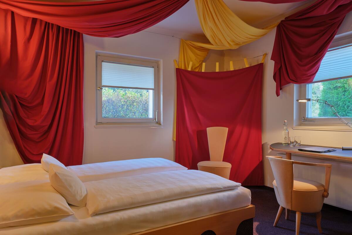 A hotel room with a bed, a desk, and a chair. There are windows with curtains, and a view of greenery outside.