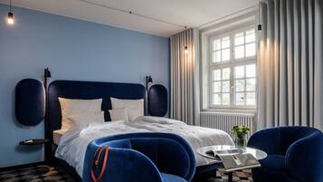 A modern hotel room with a large bed, two blue armchairs, a small round table, and a large window with curtains.