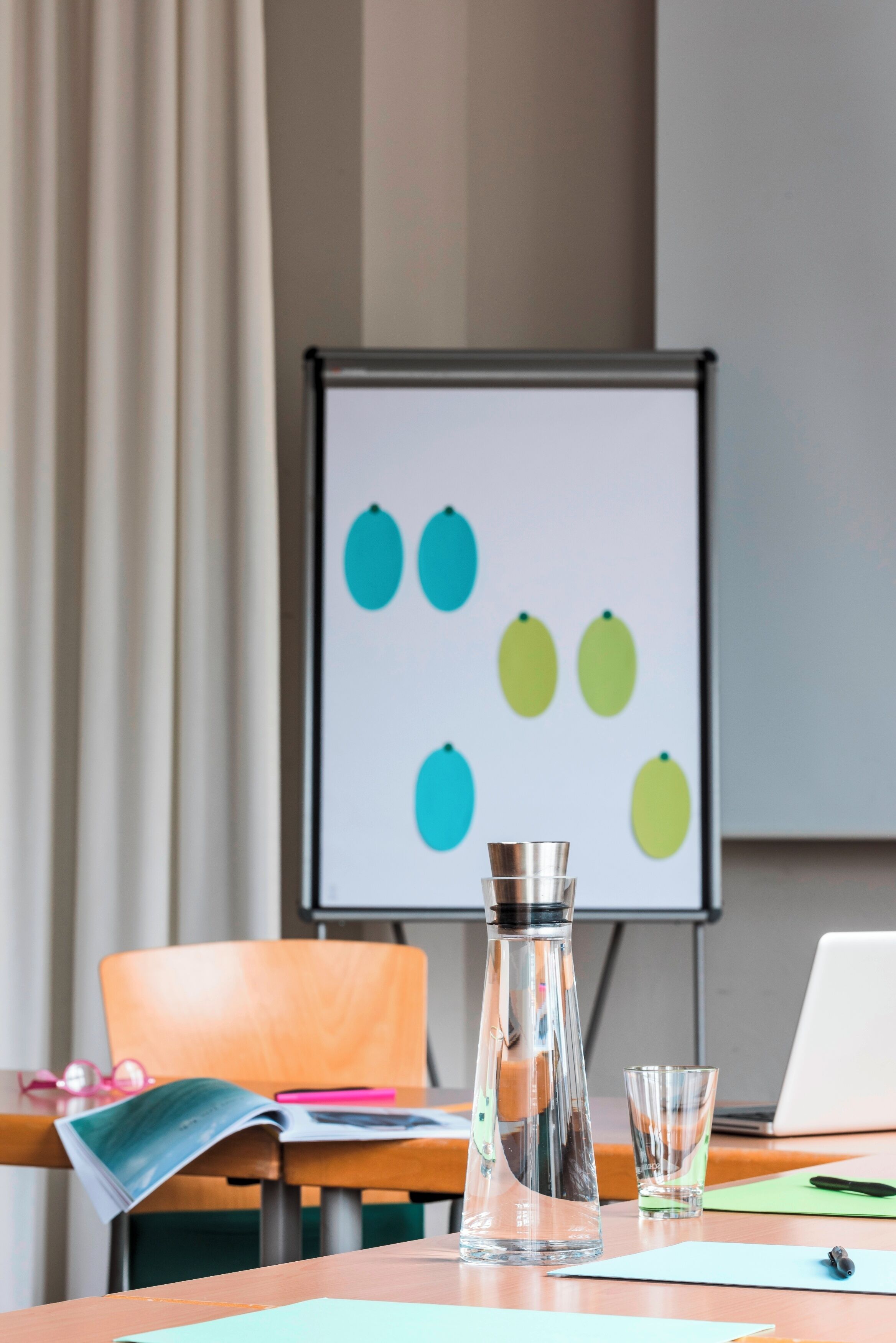 Ein Konferenzraum mit einer Tafel, auf der farbige Kreise dargestellt sind, einer Glasflasche, einem Laptop und einem Holzstuhl.
