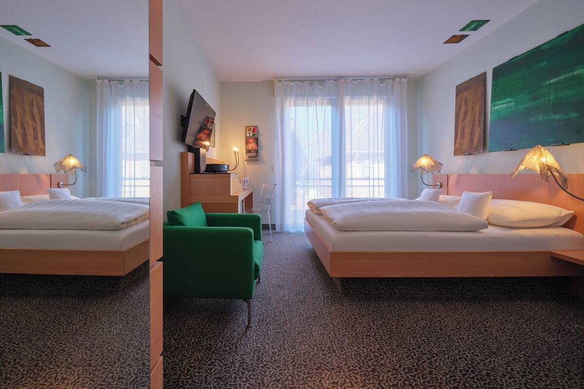 Habitación de hotel con dos camas, un sillón verde, un escritorio con silla, un televisor y obras de arte en las paredes.