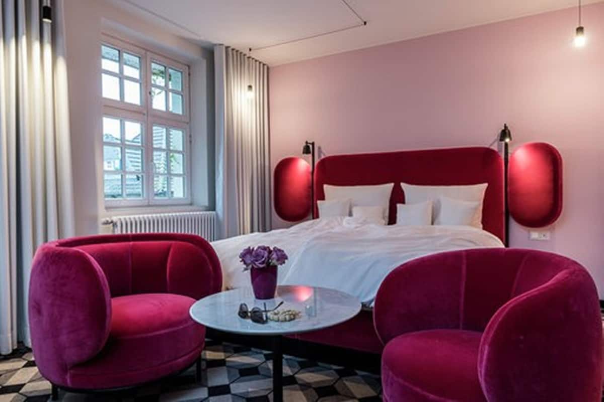Un dormitorio con una cama grande, dos sillones rojos, una mesa redonda con un florero y una ventana con cortinas.