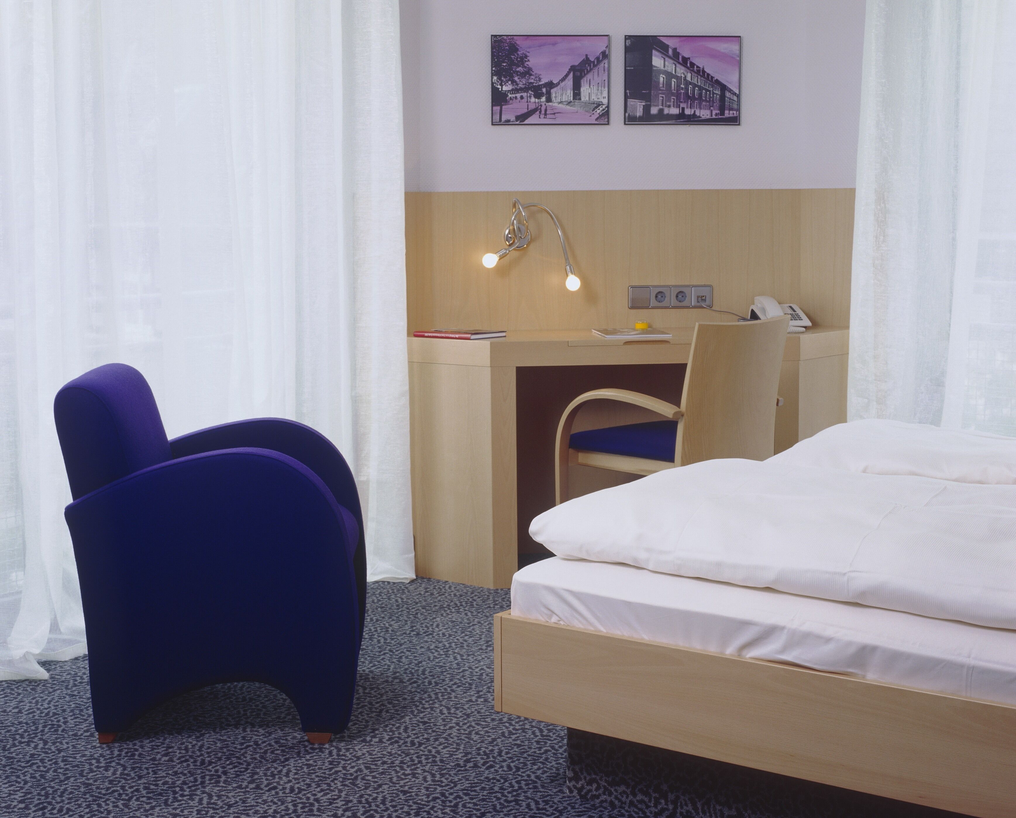A hotel room with a bed, a desk with a chair, a lamp, and two framed pictures on the wall.