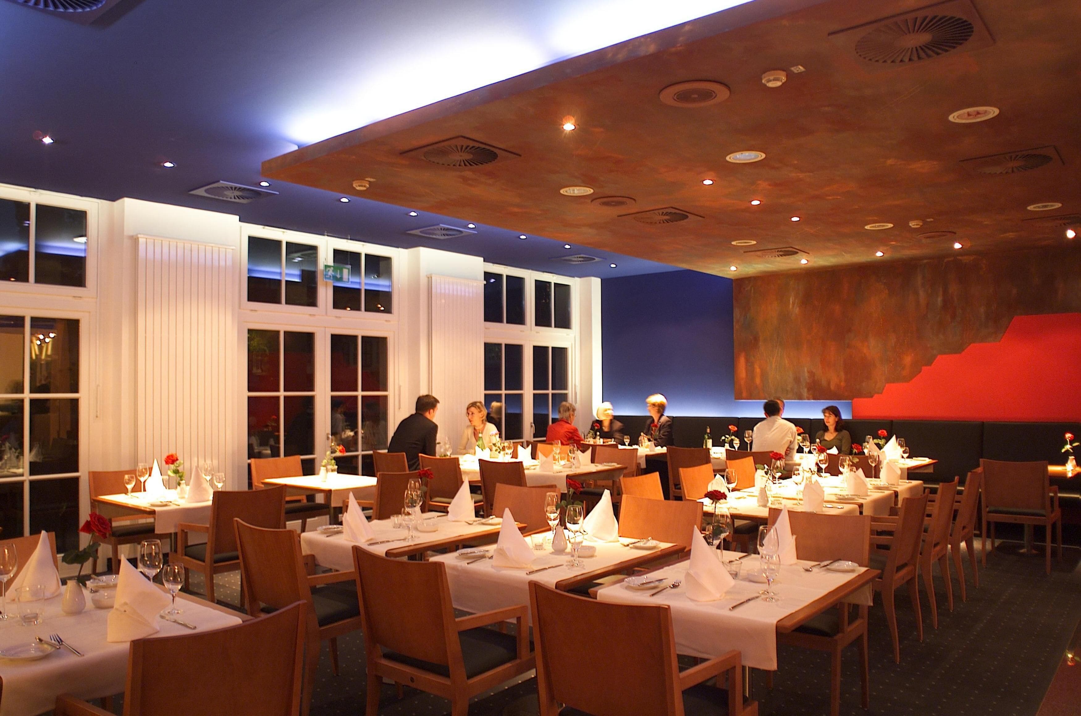 Un restaurante con varias mesas dispuestas para comer, que destaca por un diseño moderno del techo y amplios ventanales.