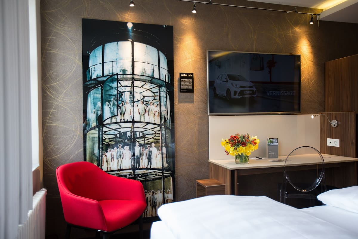 Una habitación de hotel con una cama, una silla roja, un escritorio con un florero y un cartel grande en la pared, además de un espejo que refleja un coche.