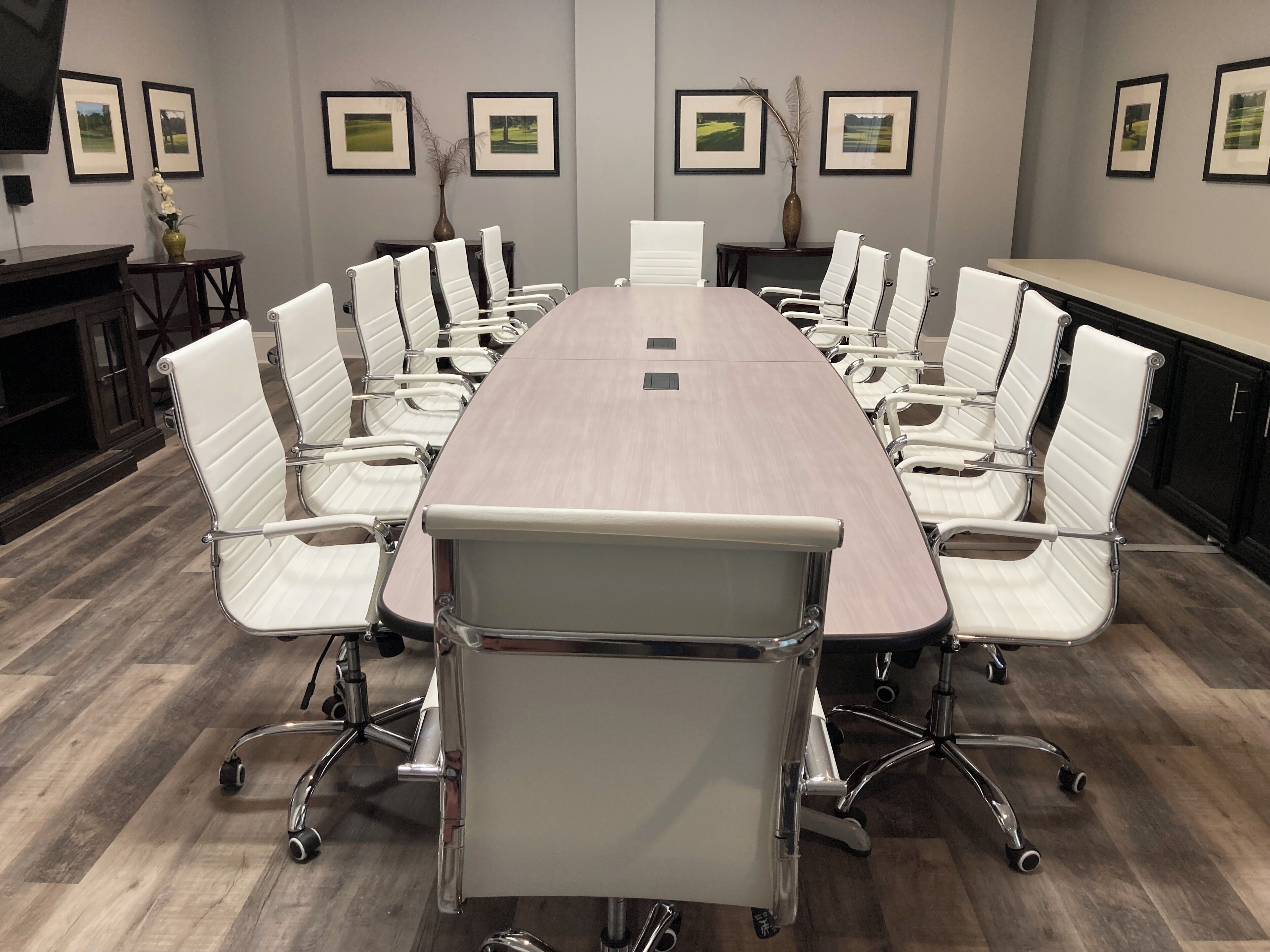 Une salle de conférence avec une longue table et des chaises blanches, des tableaux encadrés aux murs et un vase rempli de fleurs blanches.