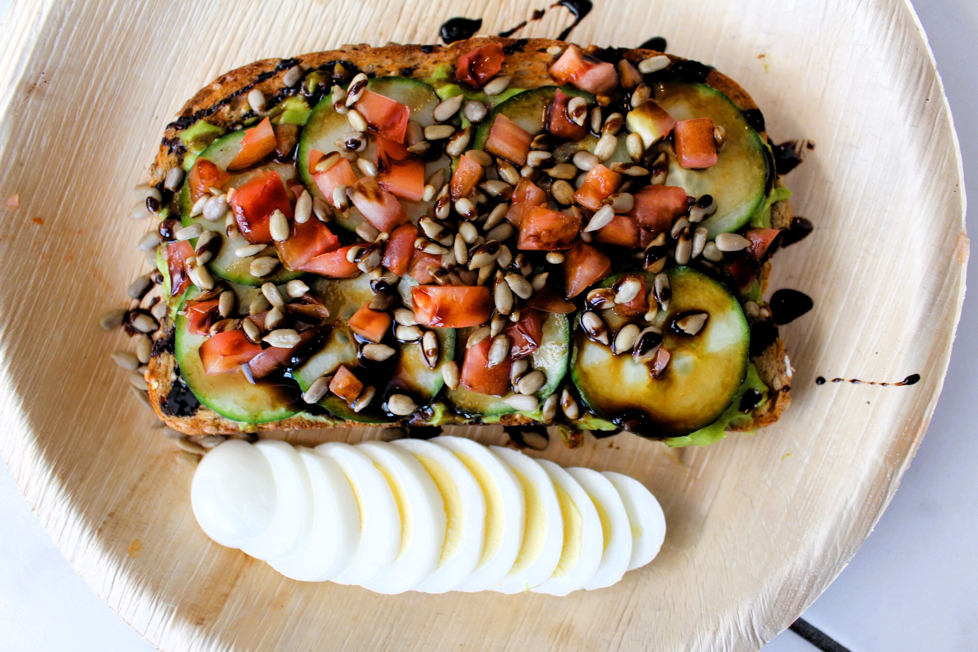 A wooden serving board with a sandwich topped with cucumber, tomato, and pine nuts, and a side of sliced white cheese.