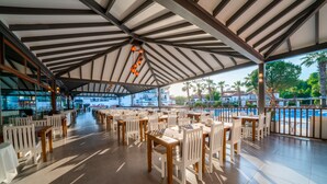 Un amplio espacio exterior para comer con varias mesas y sillas, una zona cubierta con techo a rayas, y vistas a una piscina y palmeras.