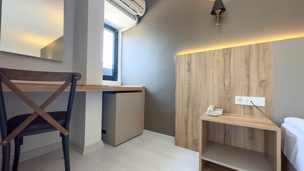 A modern, compact room with a desk, chair, and a small bedside table with a phone.