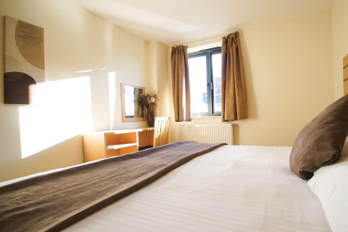 A hotel room with a bed, a desk, a chair, a window with curtains, and a radiator.