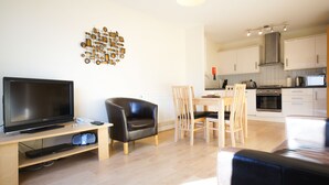 A modern living room with a flat-screen TV, a leather chair, and a dining area with wooden chairs. A kitchen with white cabinets and stainless steel appliances is visible in the background. - Crompton Court (London)