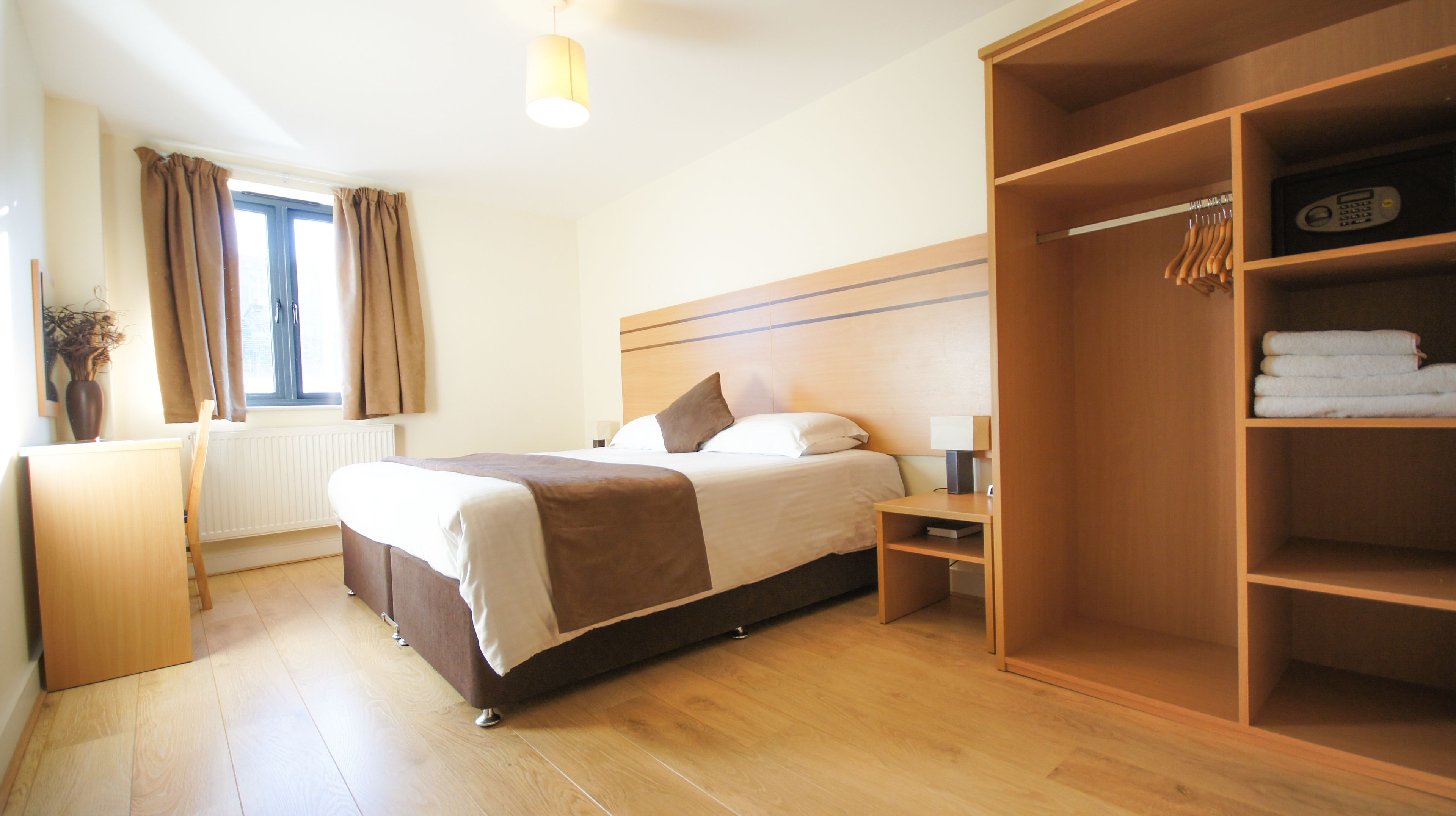 Ein Hotelzimmer mit Bett, Nachttisch, Lampe, Kleiderschrank und einem fenster mit Vorhängen.